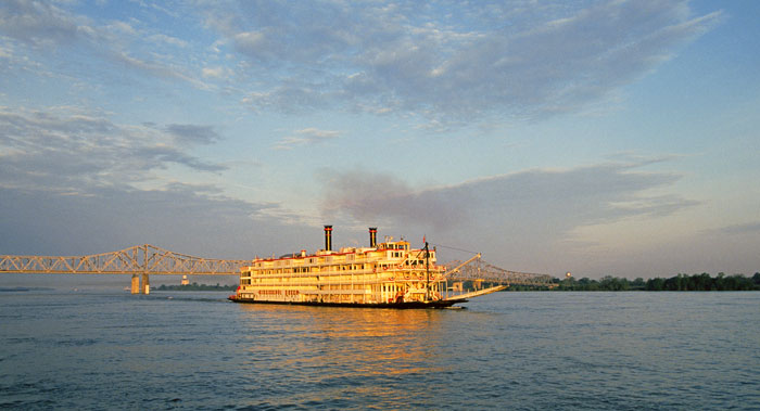 A paddlewheel steamboat cruises down the Mississippi River at sunset with a bridge in background - cruise trends to try