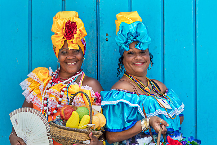 Local Cuban women in colourful traditional dress smile at the camera as they hold baskets of fruit - cruise trends to try.