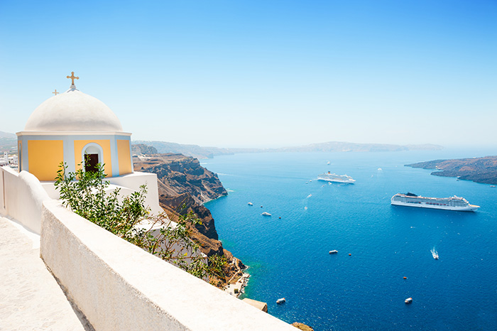 A yellow and white domed church sits on a sheer cliff overlooking passing cruise ships in the ocean below - cruise trends to try