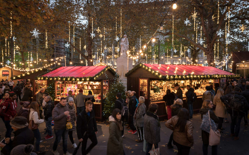 Busy markets of Christmas in Leicester Square. Fairy lights hang from a statue and the trees