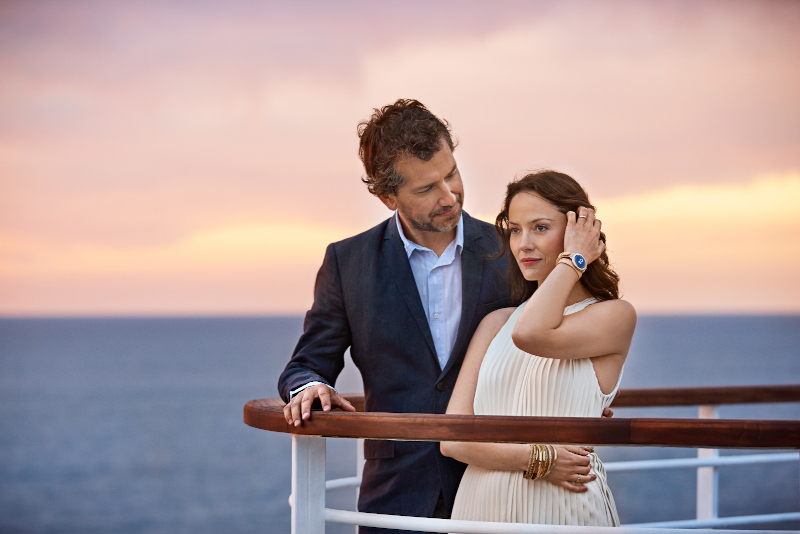 A couple standing at the back of a cruise ship as the sunsets. They are wearing the medallions