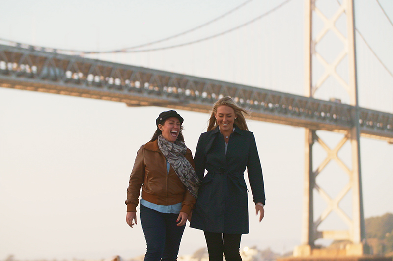 Two women walk under the shadow of the Golden Gate Bridge.