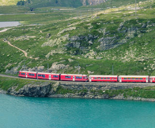 Red train going past a lake and green mountains